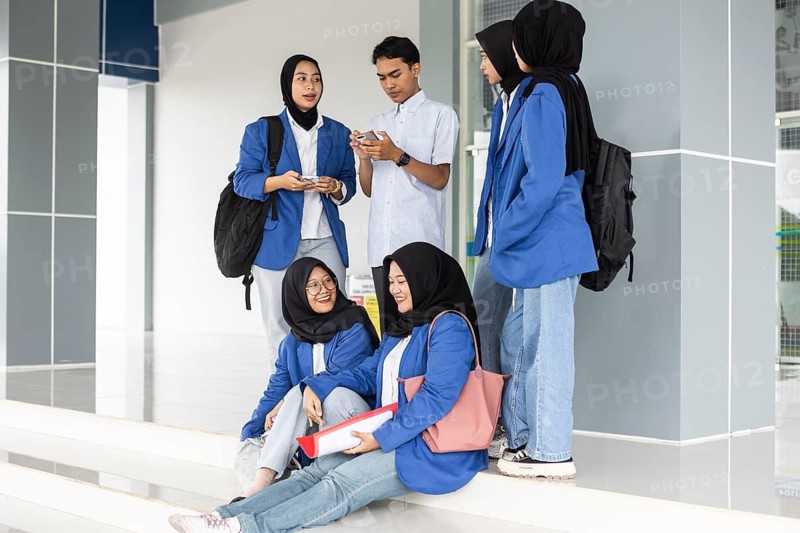 A group of Asian Muslim students gather at the school entrance, dressed in uniforms and carrying backpacks, sharing vibrant smiles and conversations as they prepare for class