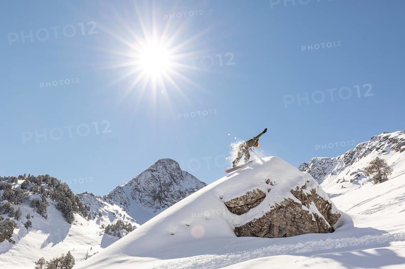 A snowboarder carves through untouched powder in a breathtaking backcountry mountain landscape. The sun shines brightly overhead, emphasizing the thrill of freeride snow sports