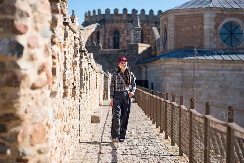 An Asian woman in casual attire walks along the cobbled medieval walls of Avila, Spain, surrounded by ancient architecture and scenic views in a historic European city