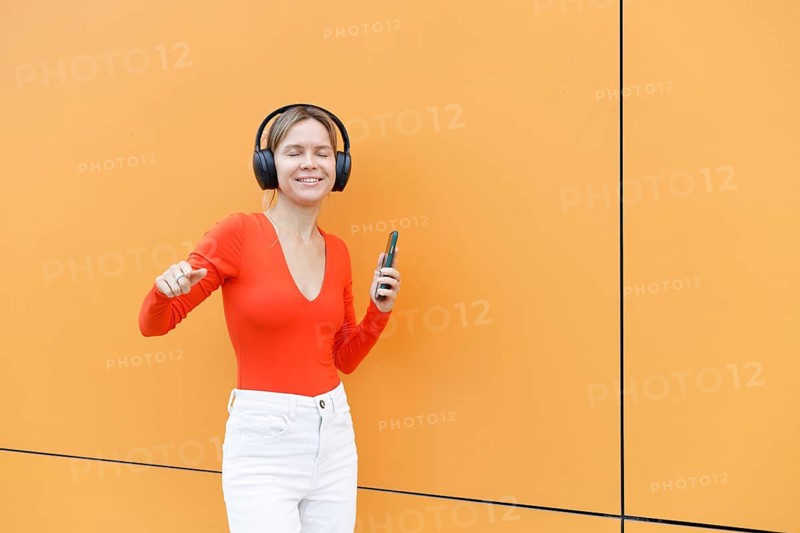 Woman in Warsaw enjoying music with headphones and smartphone She stands against a vibrant orange background, wearing a red top and white pants, smiling with her eyes closed