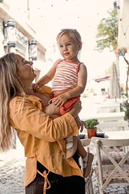A mother lovingly holds her young daughter in an outdoor setting. The sun is shining, creating a warm atmosphere as they enjoy a moment together in a quaint, charming street