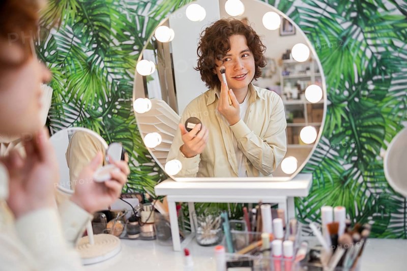 A young man is applying organic skin care with a brush in front of a lighted mirror The background features a tropical leaf pattern, and various cosmetics are on the table