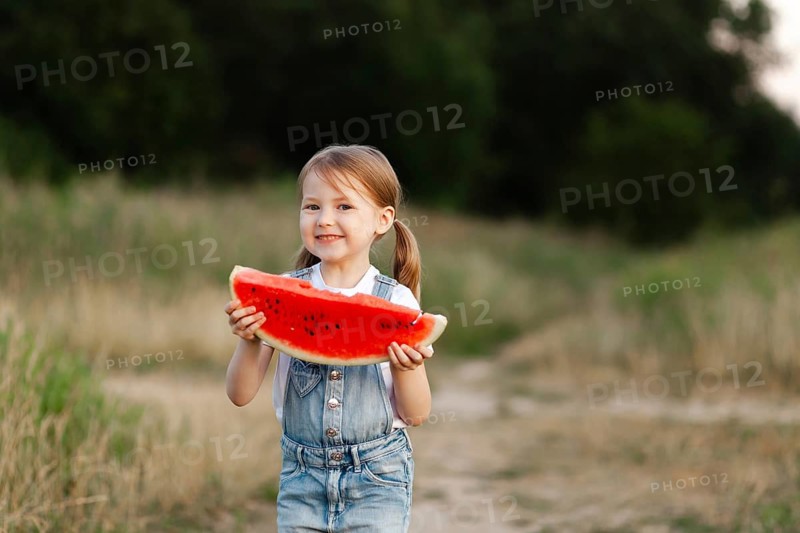 A cheerful child holds a large slice of juicy watermelon, standing outdoors in a lush green field. The warm summer day perfectly complements the refreshing fruit