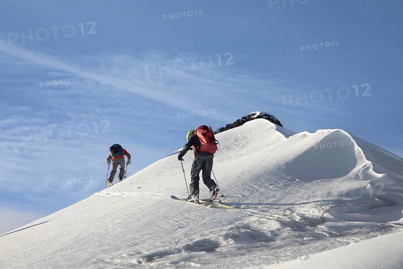 Two ski tourers traverse a pristine snow-covered mountain under a clear blue sky. Equipped with backpacks and poles, they navigate the slope with determination and skill
