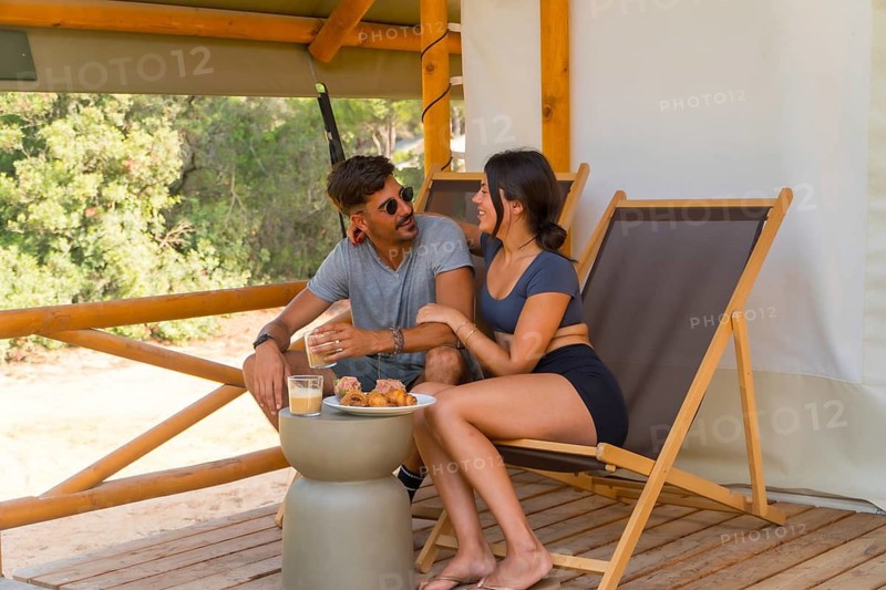 Young couple enjoying a leisurely breakfast and sipping coffee on the terrace of their glamping tent, surrounded by the beauty of nature and embracing a peaceful morning together
