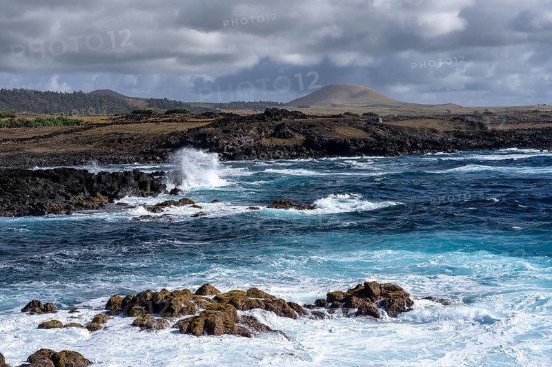 Rocky lava coast with surf and spray, volcanic cones under cloudy sky in the background, Easter Island, Chile