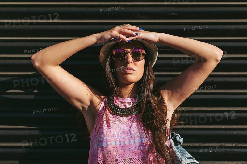 Stylish woman posing outdoors wearing a summer hat and sunglasses. Her colorful outfit and accessories highlight a trendy, carefree vibe under the sunny sky