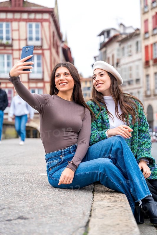 Vertical photo of two stylish friends taking a selfie sitting in a square in the city
