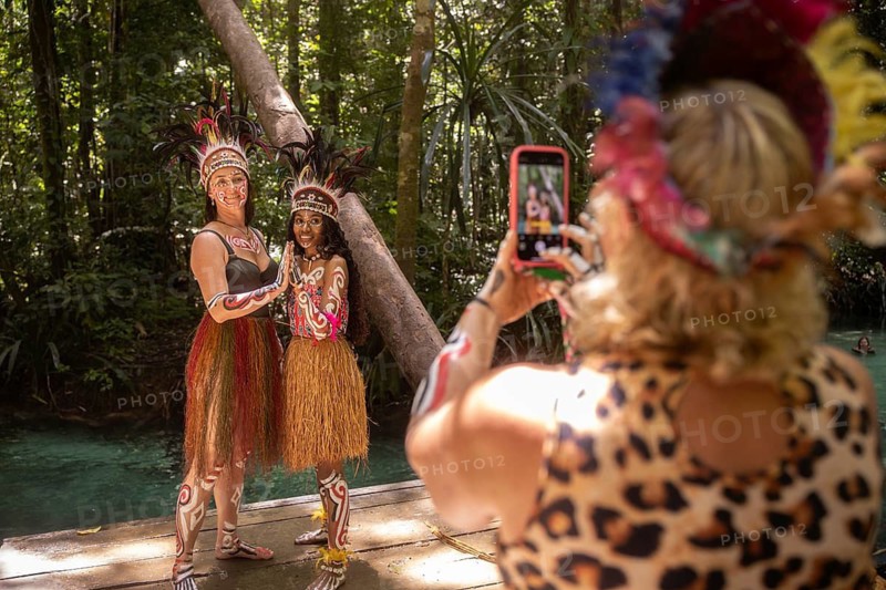 Tourist takes a photo of a friend posing with a Pacific Islander in traditional dress, deep in the tropical rainforest of Waigeo Island, surrounded by lush nature