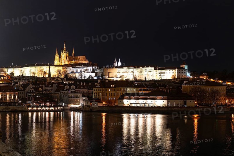 Captivating night view of Prague Castle glowing under lights, with historic architecture silhouetted against the sky. The tranquil river reflects the enchanting scenery
