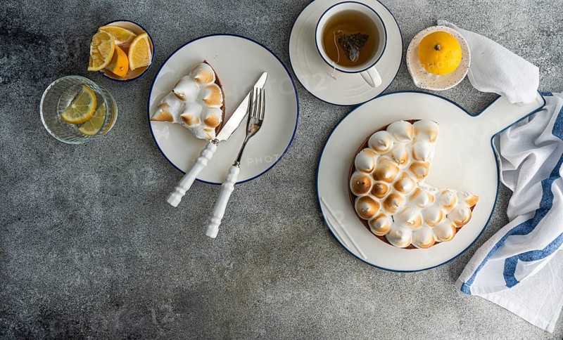 A delectable lemon meringue pie placed on a plate with a slice served alongside a cup of herbal tea. Fresh lemon slices in a glass add a refreshing touch to this cozy setting