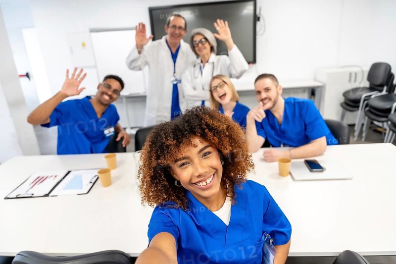 Diverse group of smiling healthcare professionals and doctors in scrubs and lab coats taking a selfie together in a modern hospital conference room, representing teamwork and unity
