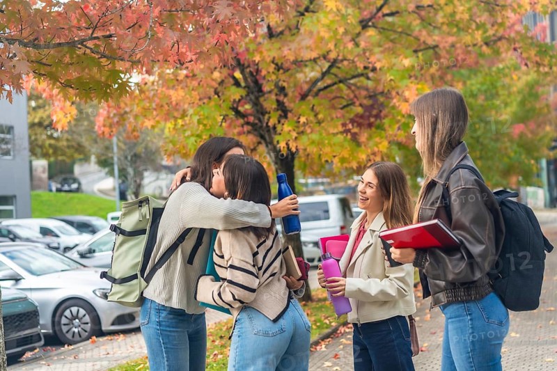 Young female students embracing and laughing on a university campus amid colorful autumn leaves, holding books and water bottles, celebrating friendship and college life
