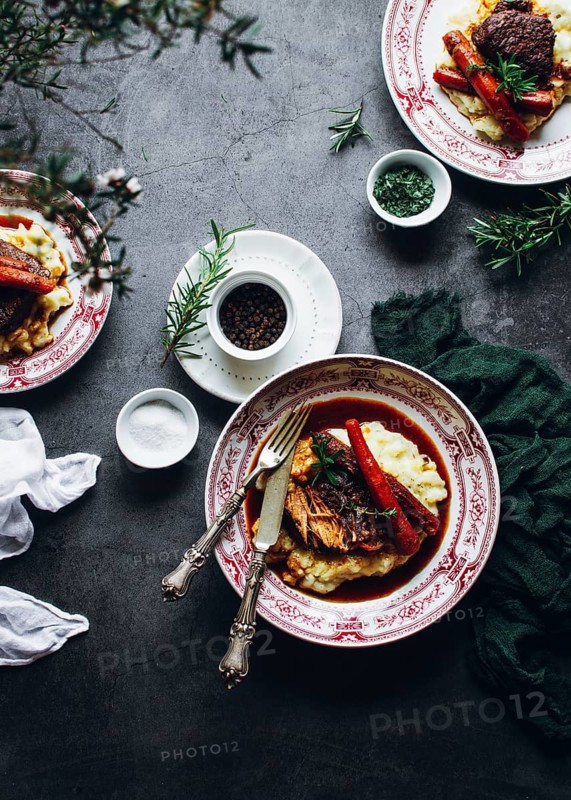 A beautifully presented rustic meal featuring mashed potatoes topped with tender meat and vegetables, garnished with fresh herbs. Elegant cutlery and spices complete the setting