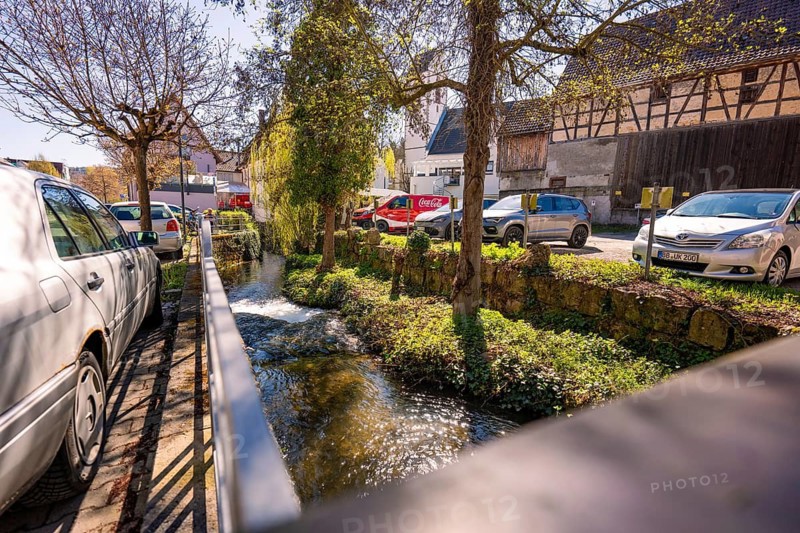 Bach flows along a road with parked cars and houses in a village, Aidlingen, Böblingen district, Germany