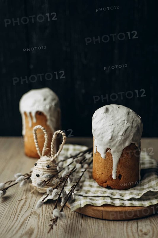 A rustic setup featuring two Kulich, traditional Russian Easter breads, generously topped with white icing. Accompanied by an Easter-themed decorative rabbit and pussy willow catkin branches, all set against a dark wooden background