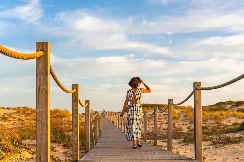 Woman in a long dress walking along a wooden boardwalk path through coastal sand dunes, enjoying a relaxing holiday evening at sunset with a clear blue sky