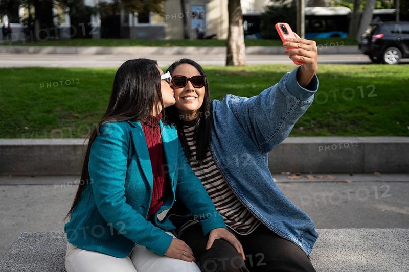 A loving latin lesbian couple sits on a park bench, capturing a joyful selfie. One partner, wearing sunglasses, holds the phone while the other lovingly kisses her on the cheek