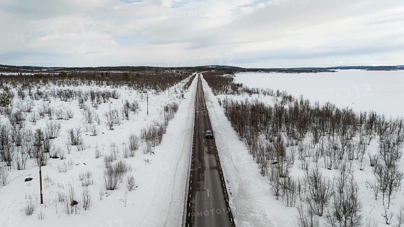 A stunning aerial view captures a long, straight road cutting through a snow-covered nordic landscape, flanked by barren trees, under a cloudy sky in a serene winter scene, Kiruna, Norrbotten, Sweden