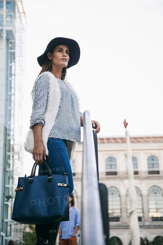 Stylish woman in jeans, sweater, and hat holding a blue handbag, posed thoughtfully in an urban environment. Reflects urban fashion and contemporary style