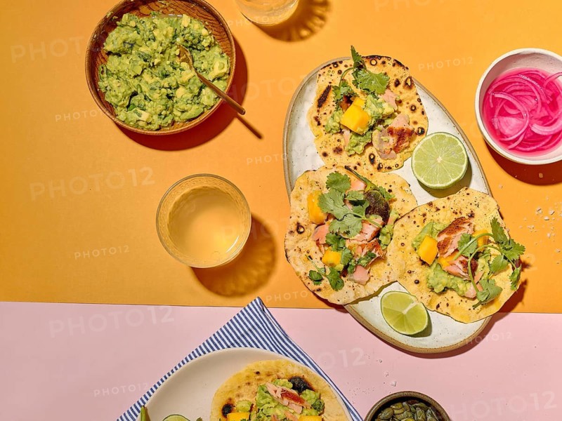 From above, vibrant taco ingredients are displayed on a yellow and pink background. The plate features tacos with mango, coriander, avocado, lime, and seasoned fish, accompanied by sides of guacamole, pickled onion, and a glass of beverage