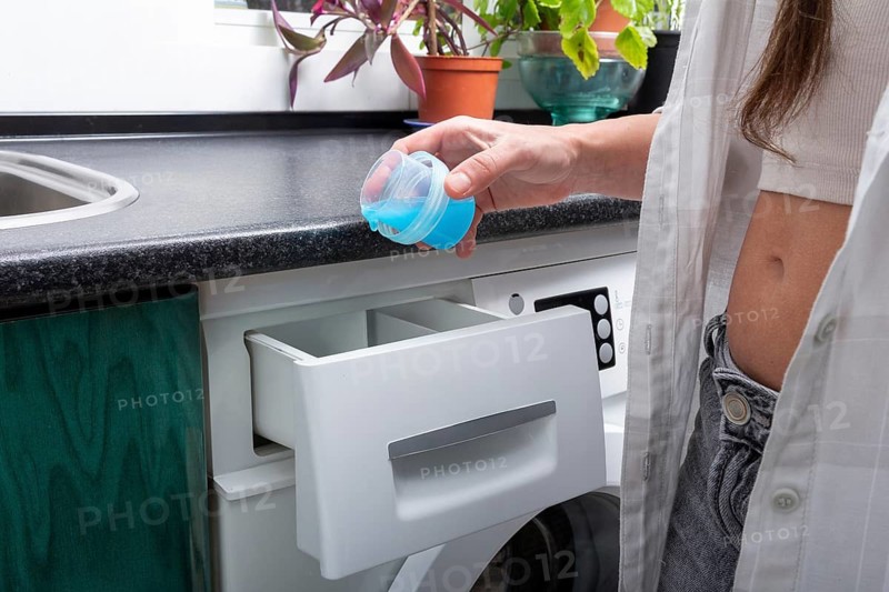A young woman stands at a front-loading washing machine, holding a blue detergent cap ready to pour, surrounded by a neat laundry space with plants and natural light