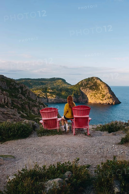 Back view of an unrecognizable woman sitting on pink chairs, overlooking the lush cliffs and Atlantic Ocean from Signal Hill, St. John's, Newfoundland & Labrador, Canada. This peaceful scene captures a moment of solitude amidst nature's beauty