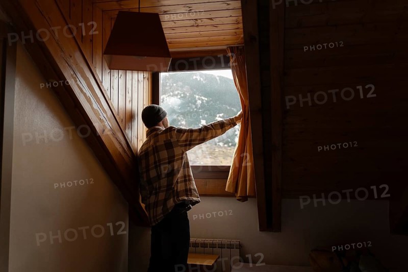 A man in a plaid shirt opens a curtain inside a cozy wooden cabin, revealing a picturesque snowy mountain landscape outside, embodying a serene winter morning