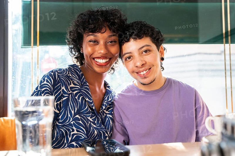 A smiling multiethnic LGBTQ+ couple enjoys a relaxed moment at a cozy café. They sit closely together at a table, radiating warmth and happiness