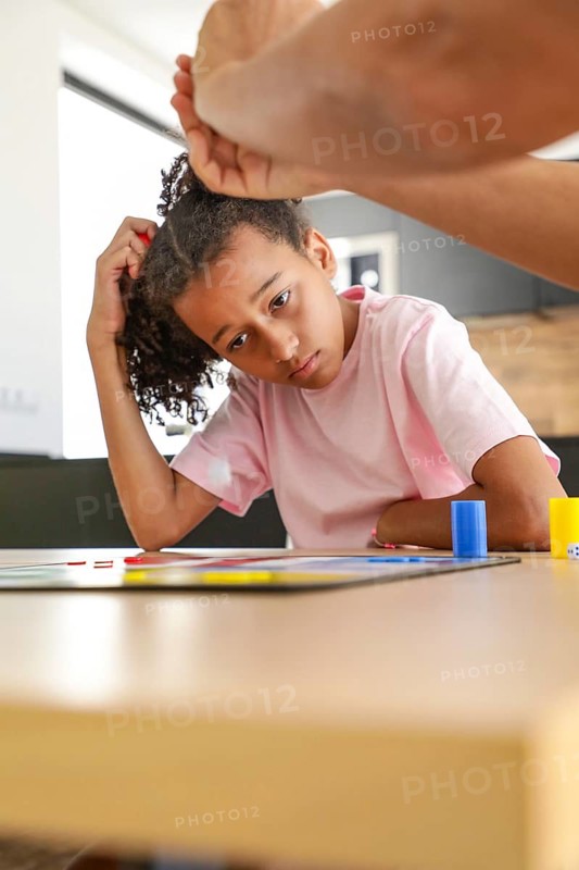 A young child, deep in thought, plays a board game at home. Surrounded by family, they focus intently on the game, enjoying a cozy day indoors together