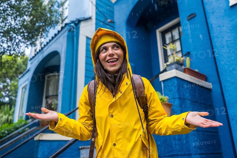 Woman wearing a yellow raincoat and beanie smiling while standing in the rain, hands open to catch raindrops, with a vibrant blue building facade in san francisco as the background