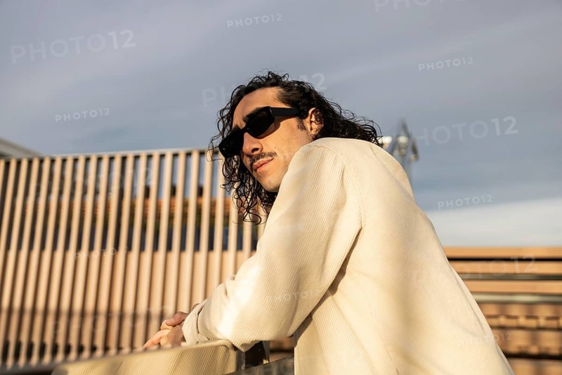 A fashionable man with long hair and sunglasses leans casually against a railing He is dressed in a light-colored shirt, and the background features a modern urban scene