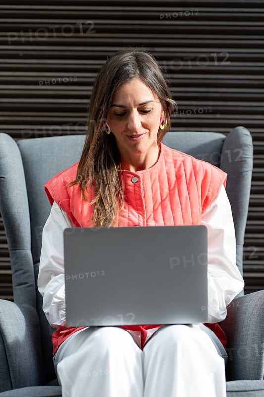 A fashion woman wearing a red and white outfit focuses on her laptop, sitting in a modern armchair. She is engaged in her work, reflecting concentration and professionalism