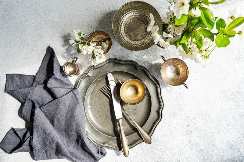 A top-down view of a table set with metal dishes, cutlery, and a gray linen napkin. Rustic elegance enhanced by delicate white blossoms in the decor