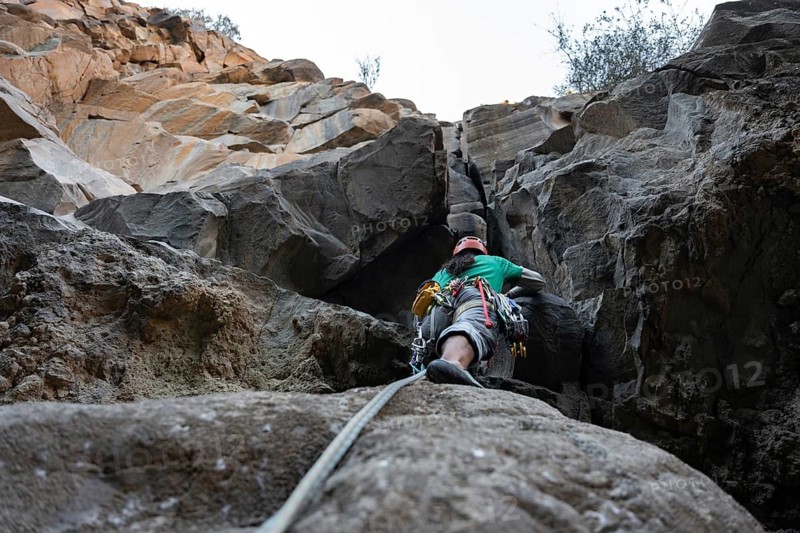 A climber in harness and helmet navigates a challenging natural rock face, displaying skill and determination. The rugged landscape emphasizes the thrill of traditional climbing