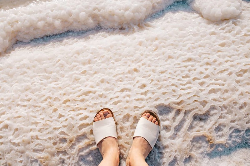 Cropped unrecognizable view of a person's feet in white sandals standing on the textured salt deposits of the Dead Sea in Jordan, highlighting a unique natural phenomenon