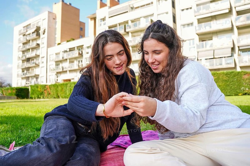 A loving LGBTQ+ young women couple sits on a picnic blanket in a grassy urban park, smiling as they affectionately engage with each other, with apartment buildings in the background