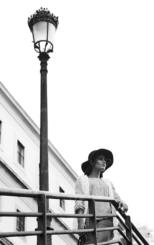 Black and white photo of a fashionable woman in a hat standing near an ornate street lamp with an urban building in the background, creating a classic urban scene