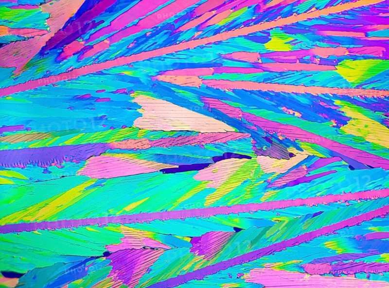 A detailed microscopic image showcases the stunning and colorful crystalline patterns of adipic acid. Vivid blues, pinks, greens, and purples dominate the palette, creating an abstract and artistic visual