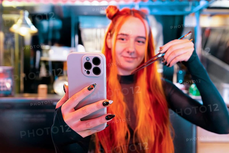 A woman with vibrant hair takes a selfie while visiting an Asian food market, holding chopsticks playfully The scene is colorful and lively, capturing the essence of travel and cultural immersion