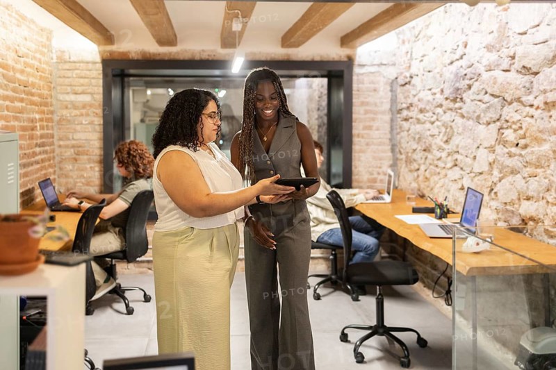 A diverse group of professionals women collaborates in a trendy coworking space, utilizing technology for productive discussions. Laptops and tablets aid in their creative work processes