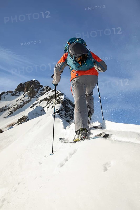 A skier wearing gear ascends a snowy mountain with ski poles, surrounded by clear skies and rugged peaks. A perfect depiction of winter adventure and exploration