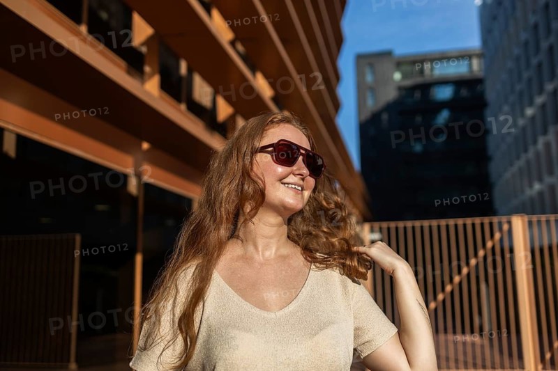A cheerful woman with long red hair and sunglasses smiles warmly, basking in the sun. Her relaxed demeanor radiates confidence, embodying a carefree urban lifestyle