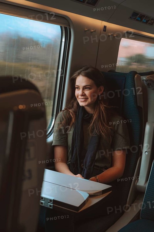 A woman smiles warmly while sitting by a window on a European train. She enjoys the passing scenery, reflecting the joy of travel and exploration across a beautiful landscape