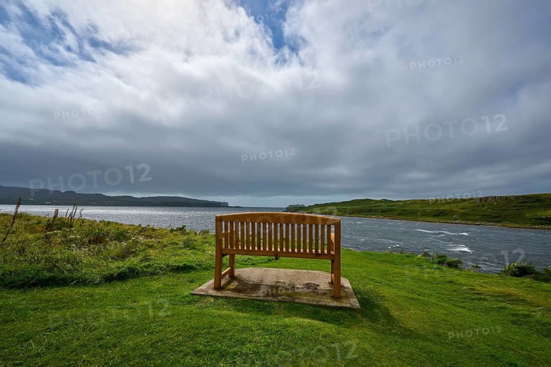 Wooden bench on shore surrounded by green landscape under cloudy sky, Isle of Skye, Scotland, United Kingdom, Great Britain