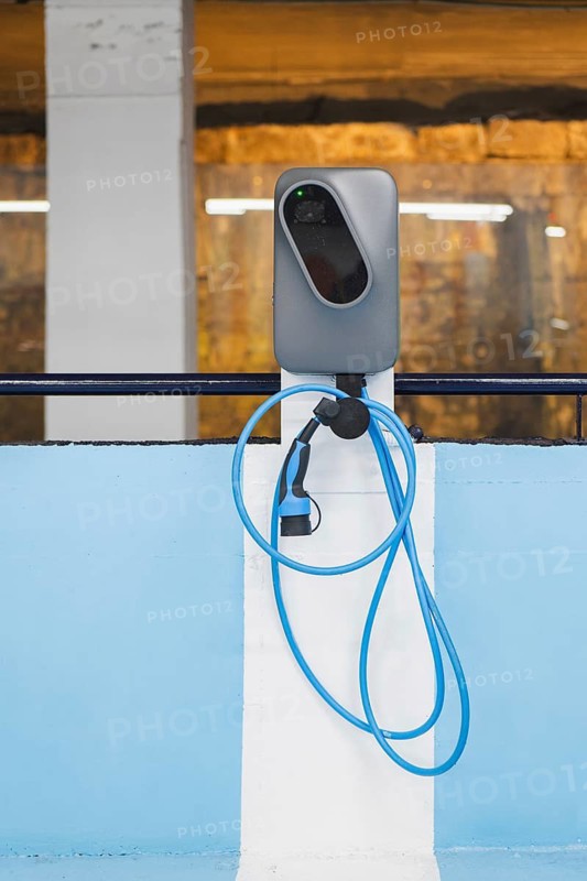 A modern electric vehicle (EV) charging station, equipped with a blue charging cable, set against a textured urban background. The station provides eco-friendly charging options