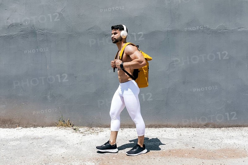 Fit man in white leggings and headphones walking confidently along a textured gray wall, carrying a bright yellow backpack. Demonstrating a sporty, urban lifestyle