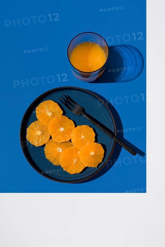 A minimalist setup featuring sliced oranges on a blue plate with a dark fork, accompanied by a glass of orange juice on a vibrant blue background