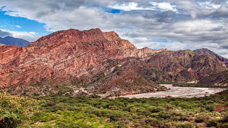 A valley with red rocks and vegetation under a cloudy sky surrounded by mountains, the landscape and colorful rock formations of the Quebrada de Cafayate near Salta in Argentina