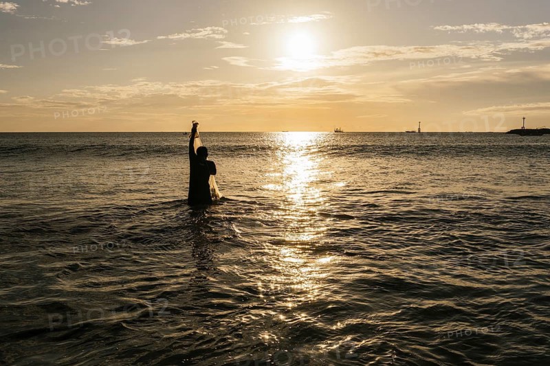 A fisherman stands in the ocean, silhouetted against the golden sunset, casting his net into the glistening water, capturing the essence of serene coastal life
